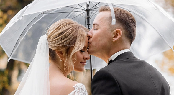 Het bruidspaar deelt een romantische kus onder een doorzichtige paraplu, een intiem huwelijksmoment in de buitenlucht.