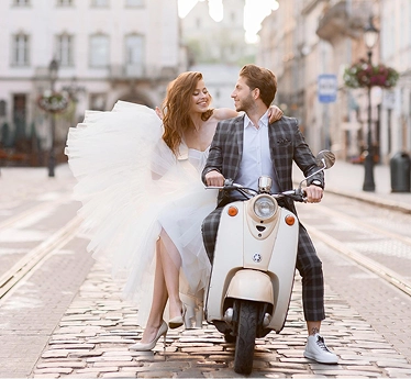 Het bruidspaar rijdt op een scooter door een straat in de stad, glimlachend en samen genietend van een romantisch huwelijksmoment.