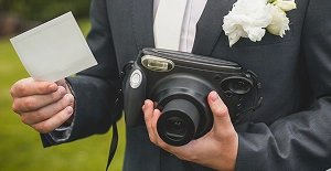 Een bruidegom in pak met een instantcamera en een afgedrukte foto in zijn handen, terwijl hij buiten spontane trouwherinneringen vastlegt.
