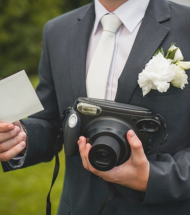 Een bruidegom in pak met een instantcamera en een afgedrukte foto in zijn handen, terwijl hij buiten spontane trouwherinneringen vastlegt.